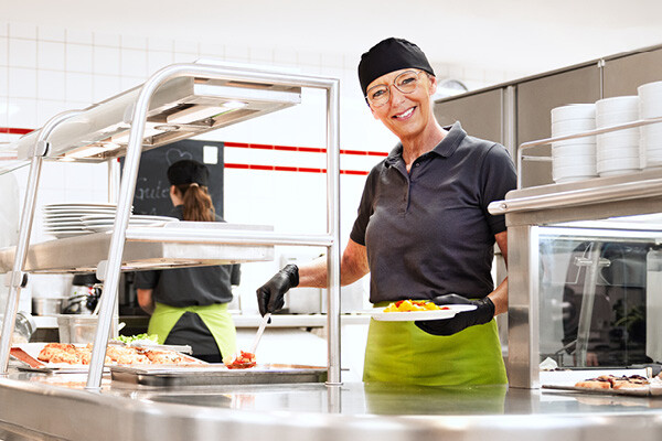 Dussmann employee from the food service and hospitality division serving food in the cafeteria and smiling.