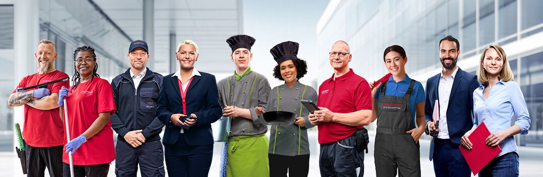 Group picture of smiling Dussmann employees from different work areas in front of a gray background, wearing their work clothes. 